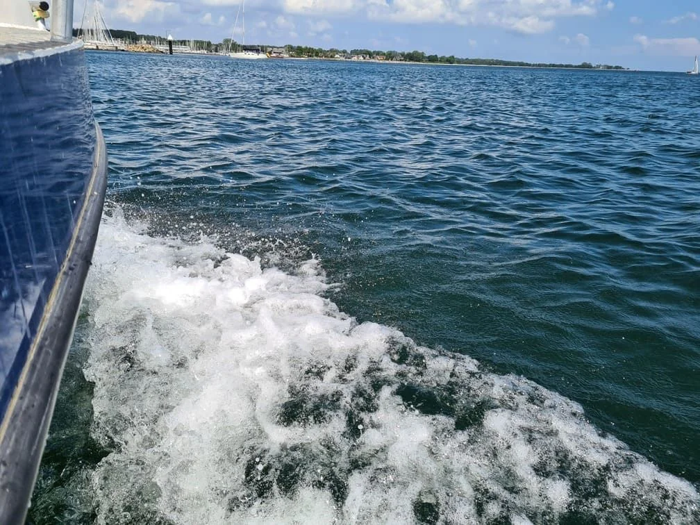Boat hull cutting through ocean waves with coastal marina and sailboats visible in background.