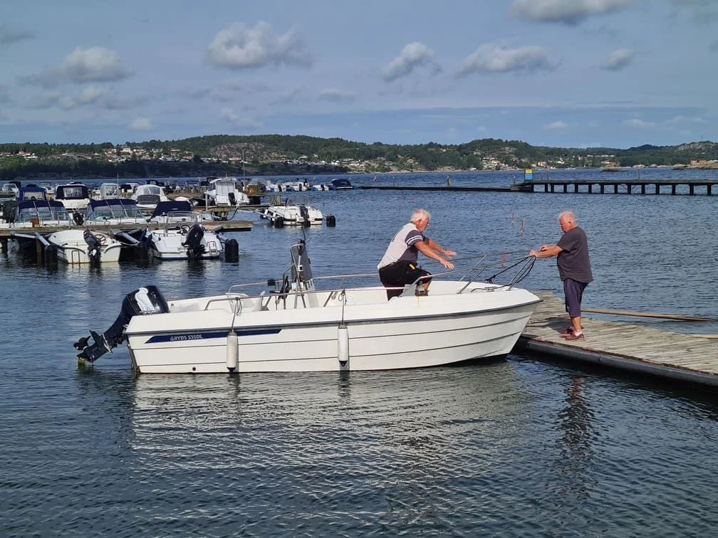 Two men inspect outboard motor on white boat docked at wooden pier, moored boats visible in background.