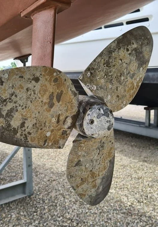 Corroded boat propeller with heavy marine growth and oxidation, mounted on shaft near dock.