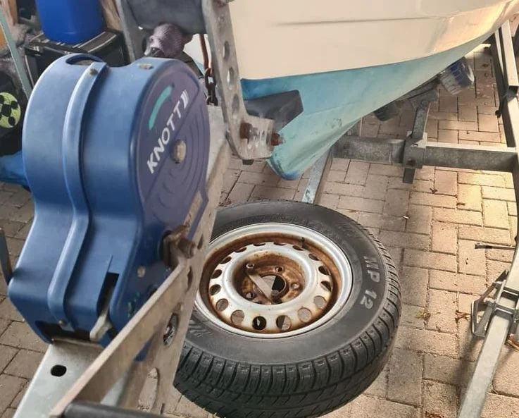 Boat trailer wheel and winch assembly on paver driveway with damaged hull visible.